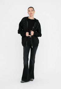 Modèle dans un sweat à capuche noir zippé orné de rivets argentés, associé à un haut noir et à un jean évasé, se tenant devant un fond blanc.