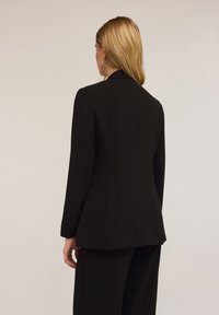 Blazer nero su misura con maniche lunghe, texture liscia, silhouette strutturata e design minimalista, visto da dietro.