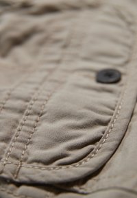 Tessuto beige con una superficie testurizzata, presenta cuciture visibili e un bottone nero su una tasca curva. La vista ravvicinata mostra i dettagli.