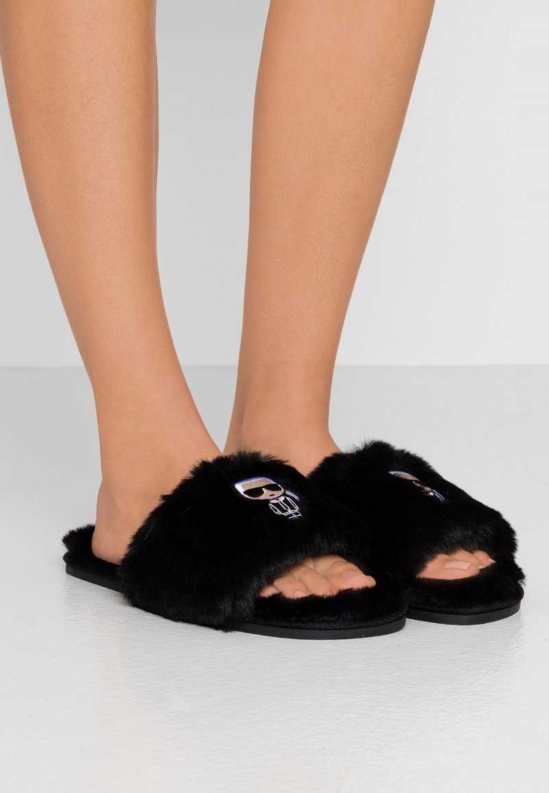 Jambes portant des chaussons noirs et fourrés à bout ouvert avec un petit motif de figure brodée sur la bande supérieure, debout sur un sol clair.