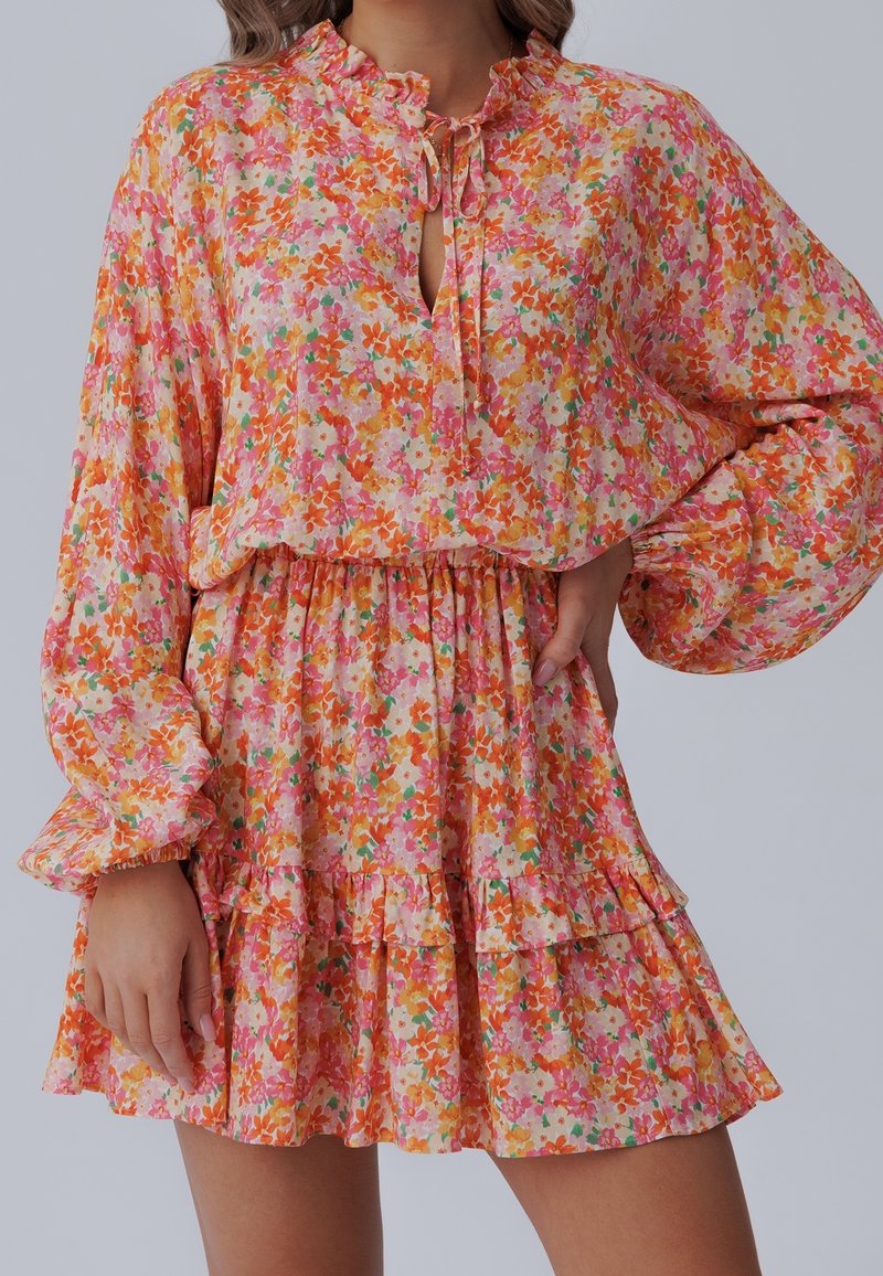 Kwiatowa mini sukienka z lekkiego materiału z wielokolorowymi wzorami w odcieniach różu, pomarańczu i zieleni, z falbanami na rękawach i na dole sukienki.