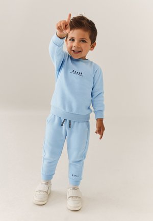 Usměvavý mladý chlapec v světle modré mikině a kalhotách ukazuje pravou rukou nahoru na jednobarevném béžovém pozadí.