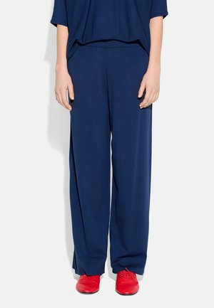 Osoba nosząca luźne ciemnoniebieskie spodnie i pasującą górę, stojąca z rozluźnionymi rękami, widoczne czerwone buty sznurowane.