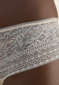 Gros plan sur de la lingerie en dentelle blanche sur une peau foncée, mettant en valeur les motifs délicats de la dentelle et sa texture.