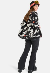 Zwart-wit fleecejack met abstract patroon en rode stippen, gecombineerd met zwarte skibroeken. Model draagt skibril en een zwarte beanie.