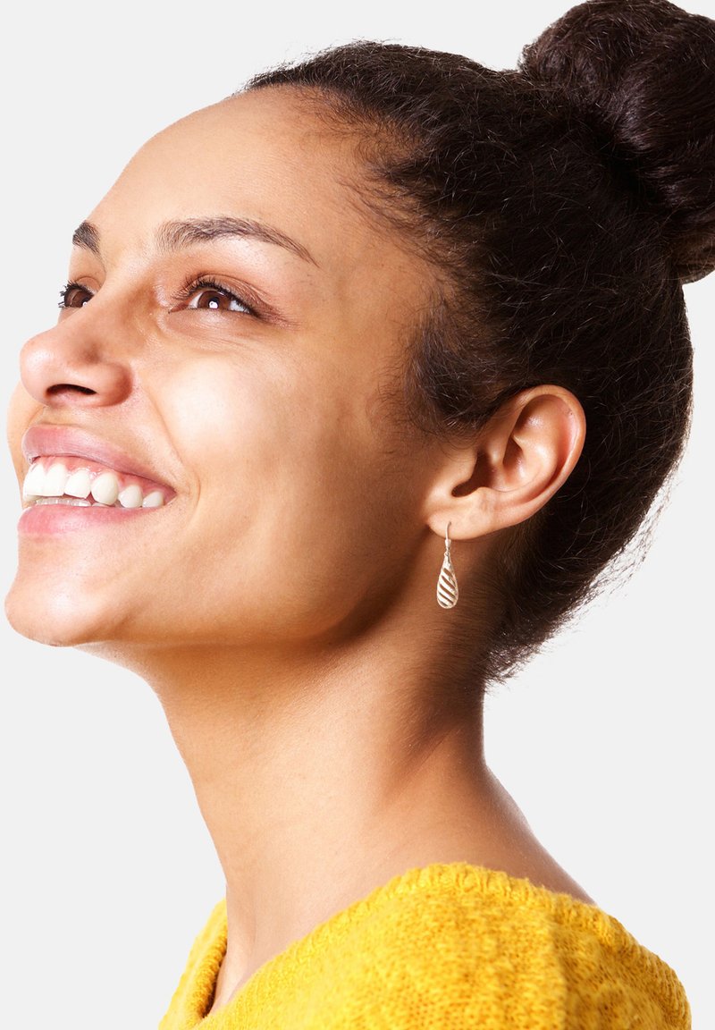 Silverhängande örhängen med en vriden, randig design, bar en person med medelbrun hy och mörkt hår, klädd i en gul tröja.