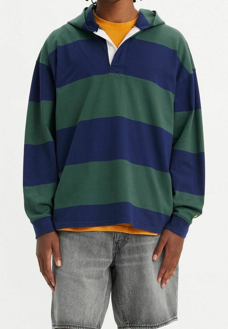 Uomo in maglietta rugby a righe oversize blu navy e verde sopra una t-shirt arancione, con pantaloni shorts in denim grigio sbiadito, in piedi contro uno sfondo semplice.