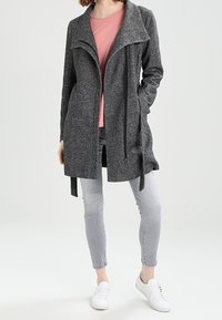 Grijze, gestructureerde lange cardigan met een open voorkant en kraag. Gepaard met een roze top, lichte jeans en witte sneakers.