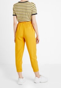 Randig gul topp med svarta accenter, kombinerad med högmidjade, korta gula byxor. Vita sneakers fulländar outfiten.