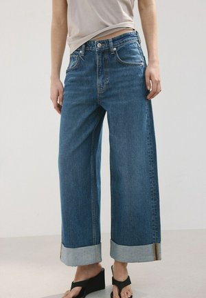 Personne portant un jean denim bleu à jambes larges avec de grands revers, associé à des sandales compensées noires, debout devant un fond uni.