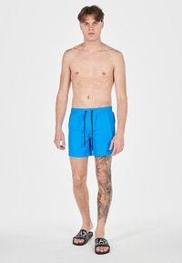 Costumi da bagno blu con vita elasticizzata, cordino regolabile e lunghezza corta. Vengono indossati sandali neri con logo bianco.