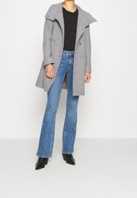 Grijze wollen blendjas met hoge kraag, dubbelknopen ontwerp en grote knopen, gecombineerd met uitlopende blauwe jeans en zwarte haklaarzen.