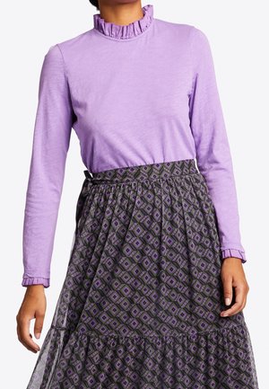 Mujer vistiendo una blusa de manga larga lavanda con cuello y puños con volantes, combinada con una falda de talle alto con estampado en tonos púrpura y gris.