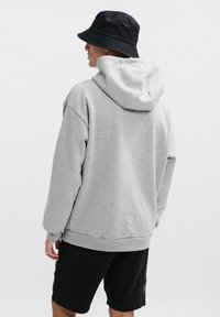 Grå fleecehuva med huva, sänkta axlar, ribbade ärmslut och nederkant. Svart bucket hat bärs, tillsammans med svarta shorts.