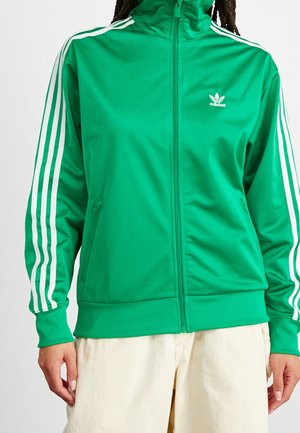 Veste zippée Adidas verte avec détails à trois bandes blanches sur les manches et logo sur la poitrine, portée avec un pantalon beige clair.