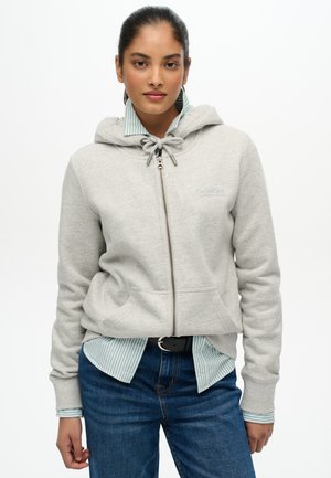 Jonge vrouw draagt een lichtgrijze hoodie met rits over een gestreept shirt en een blauwe spijkerbroek, staand tegen een effen witte achtergrond.