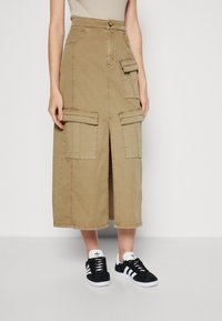 Beige midi-rok met een voorknoop en meerdere klepzakken, gedragen met zwart-witte Adidas sneakers op een lichte achtergrond.