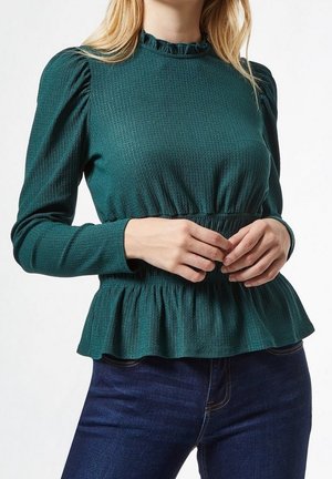 Kvinde iført en tekstureret, mørkegrøn langærmet bluse med pufskuldre og flæsekanter, kombineret med mørkeblå jeans.