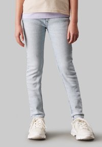 Dítě stojí oblečené v světle modrých skinny džínách, béžových a levandulových vrstvených tričkách a bílých teniskách na jednobarevném šedém pozadí.