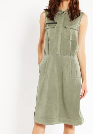 Vestito verde oliva senza maniche con colletto, cerniera frontale, due tasche sul petto e due tasche laterali indossato da una persona in piedi.