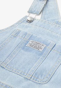 Φωτεινά γαλάζια τζιν σαλόπες με κίτρινη ραφή, με μπροστινή τσέπη και ετικέτα που γράφει "Levi Strauss & Co." στο στήθος. Ασημένια μεταλλικά εξαρτήματα.