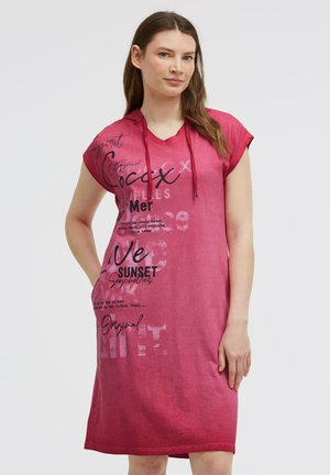 Frau mit langen braunen Haaren, die ein kurzärmeliges, rosa Kapuzenkleid mit schwarz-weißen Textgrafiken trägt, steht mit einer Hand in der Tasche.