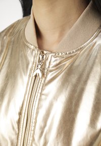 Gold Bomberjacke mit glänzendem, metallischem Finish, beigem, geripptem Kragen, Reißverschluss mit dekorativem goldenen Zip.