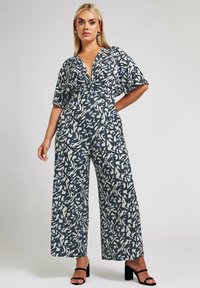 Sininen ja valkoinen kuviolaitteinen haalarimainen asu, jossa on lyhyet hihat, syvä V-pääntie ja leveät lahkeet. Kankaan pinta on sileä. Mustat lohkokorkoiset sandaalit.