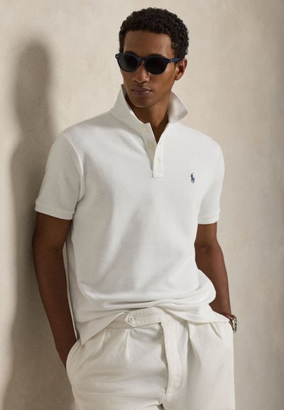 Mężczyzna w czarnych okularach przeciwsłonecznych, białej koszulce polo z podniesionym kołnierzem i białych spodniach, stojący z rękami w kieszeniach na tle beżowej ściany.