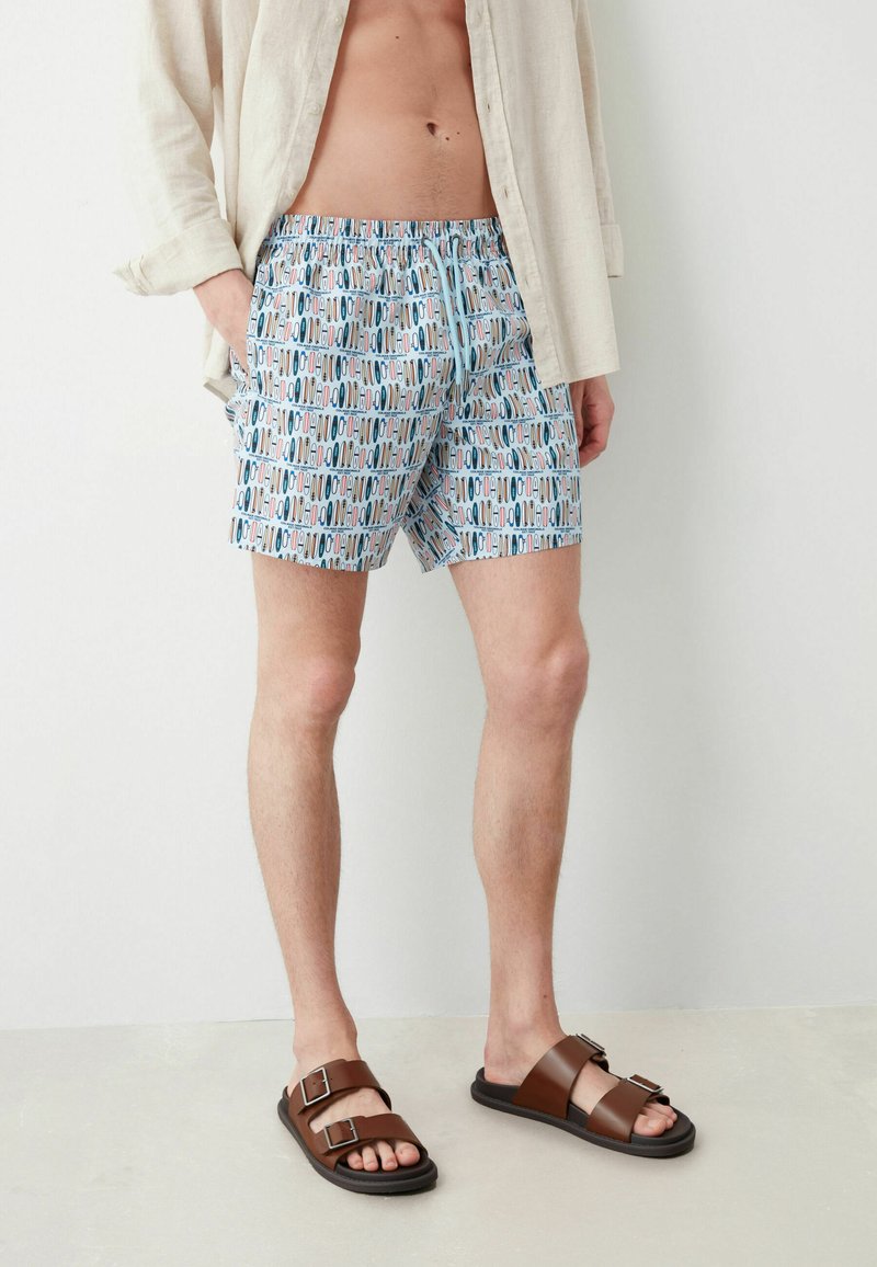 Costume da bagno in un modello blu chiaro con forme astratte multicolori. Indossato con sandali marroni a doppia fascia e una leggera camicia di lino.