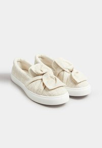 Scarpe slip-on in tessuto beige con un grande dettaglio a fiocco sulla parte superiore, suole in gomma bianca e superficie texturizzata.