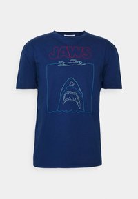 Marineblauwe katoenen T-shirt met het woord "JAWS" in het rood en omrandde graphics van een zwemmer en een haai in teal stiksel.