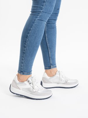 Feet wearing white and silver lace-up sneakers paired with fitted blue jeans, shown against a plain white background.