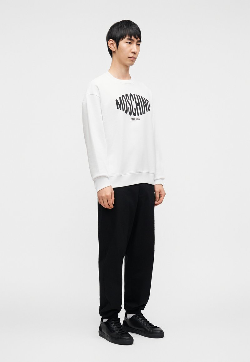 Sweat-shirt blanc avec logo "Moschino" noir, associé à un pantalon noir et des baskets noires. Le sweat-shirt présente une coupe décontractée et des poignets côtelés.