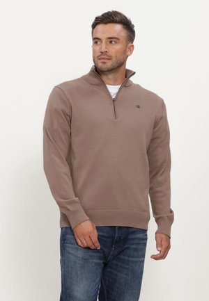 Uomo che indossa un maglione marrone con zip parziale sopra una camicia bianca e jeans blu, guarda a sinistra su uno sfondo semplice.