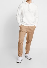 Vit hoodie med präglad "LACOSTE"-logotyp, kombinerad med beige chinos och vita sneakers med grå detaljer. Enkel design, ledig passform.