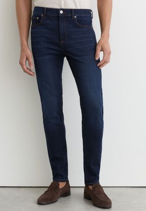 Mand iført mørkeblå slim-fit jeans med brune ruskindsløbere, stående mod en hvid væg på en grå gulv.