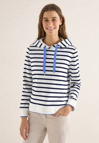 Femme souriante, portant un sweat à capuche blanc à rayures horizontales bleu marine et cordons de serrage bleus, ainsi que un pantalon beige, debout avec une main dans la poche.