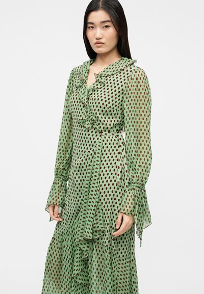 Γυναίκα με μακριά σκούρα μαλλιά, που φοράει ένα πράσινο φόρεμα τύπου wrap με καφέ πουά και βολάν, στέκεται μπροστά σε ένα απλό φωτεινό φόντο.