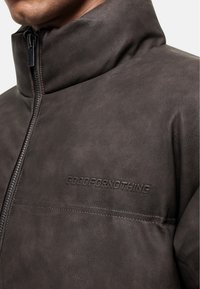 Veste en cuir synthétique marron avec un col montant, une fermeture éclair à l'avant et un logo en relief "GOODFORNOTHING" sur la poitrine, présentant une texture lisse.