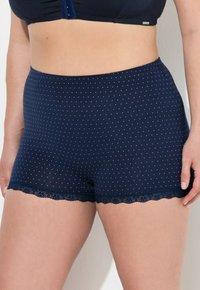 Donna che indossa una mutanda boyshort a vita alta di colore blu navy con piccoli pois bianchi e rifiniture in pizzo, tagliata a metà vita e alle cosce.