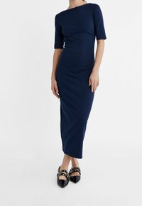 Granatowa, dopasowana sukienka midi z dekoltem typu boat i krótkimi rękawami. Noszona z czarnymi szpilkami zakończonymi w szpic, zdobionymi ćwiekami.