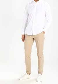 Biała koszula z długim rękawem zapinana na guziki zestawiona z beżowymi spodniami slim-fit i białymi sneakersami. Koszula ma standardowy kołnierzyk oraz gładką teksturę.