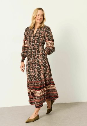 Woman with blonde hair wearing a long floral and patterned dress and olive green flats, standing against a plain light background.