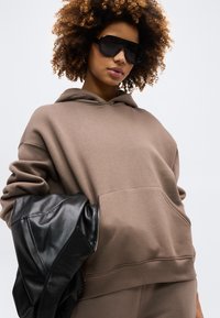 Γυναίκα με σγουρά μαλλιά, που φοράει υπερμεγέθη καφέ φούτερ με κουκούλα και μεγάλα μαύρα γυαλιά ηλίου, κρατώντας μαύρο δερμάτινο μπουφάν πάνω σε ένα μπράτσο.