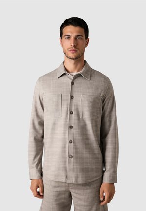 Hombre vestido con camisa de cuadros beige abotonada y pantalones a juego, de pie frente a un fondo liso gris claro, mirando hacia adelante.