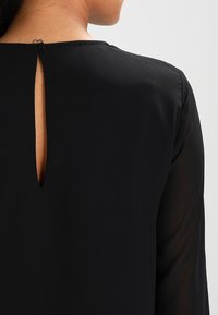 Czarna bluzka z otworem w kształcie klucza z tyłu, o gładkiej fakturze i długich rękawach, wykonana z lekkiego materiału.