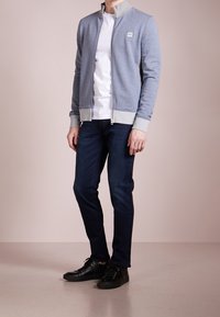 Uomo che indossa una giacca zip blu chiaro, una maglietta bianca, jeans blu scuro e sneaker nere in piedi su un pavimento beige.