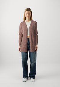 Long gilet à ouverture frontale en tricot doux rose poussiéreux. Comprend deux poches avant et des poignets à côtes. Associé à un jean taille haute déchiré.