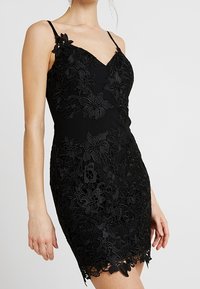 Robe noire au silhouette ajustée, ornée de dentelle à motifs floraux, bretelles fines et ourlet festonné.